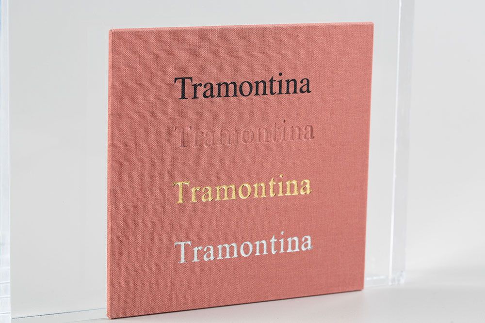 The word 'Tramontina' is displayed in four different font styles on a pink or coral-colored background.