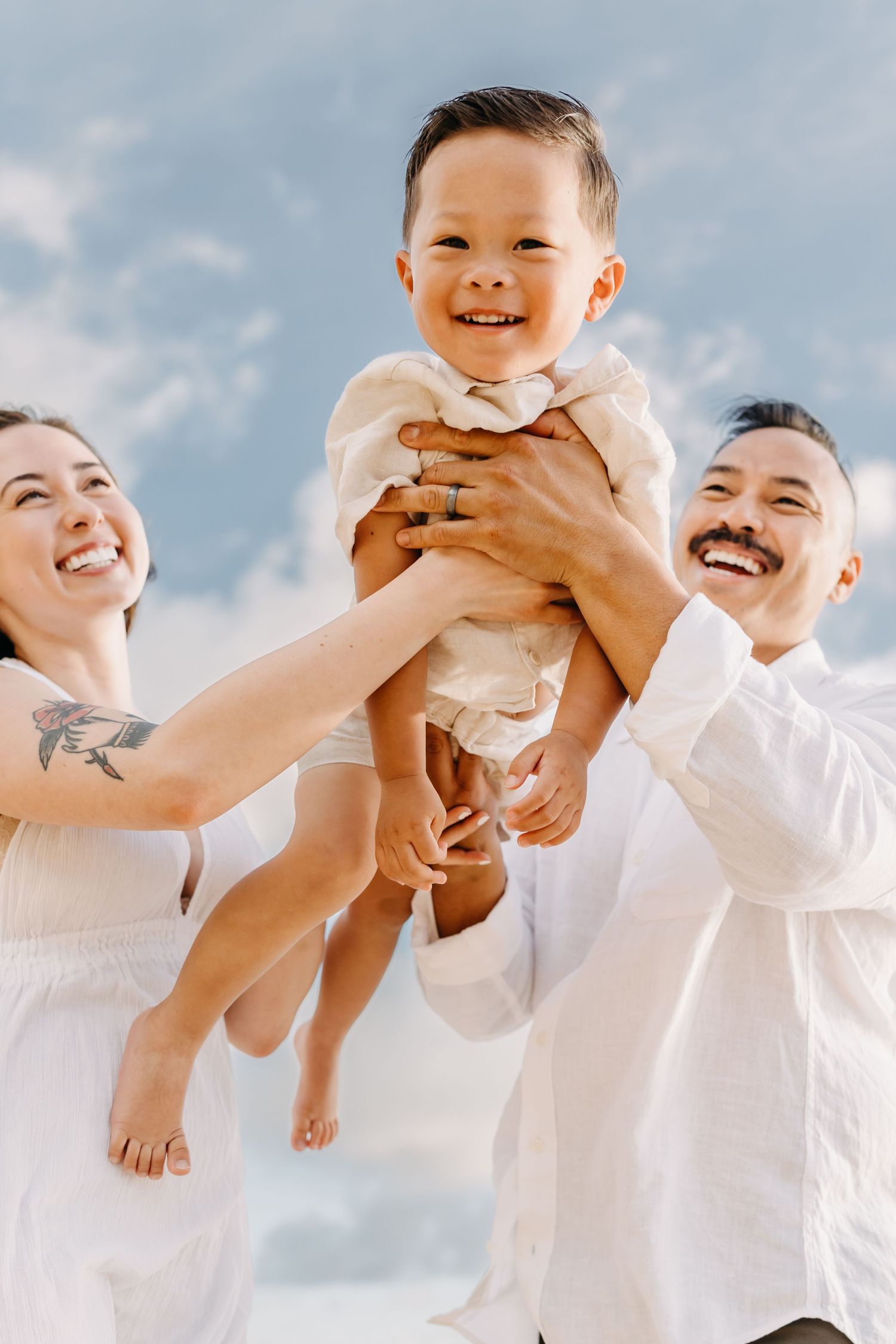 Parents in white outfits lift and play with their happy barefoot toddler against a bright blue sky background.