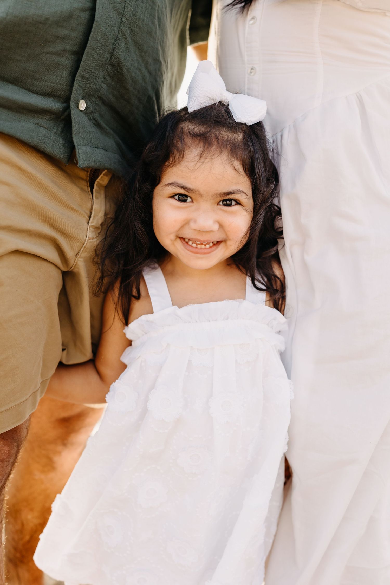 Young child wearing a white dress and bow smiles between two adults in tan and white clothing.