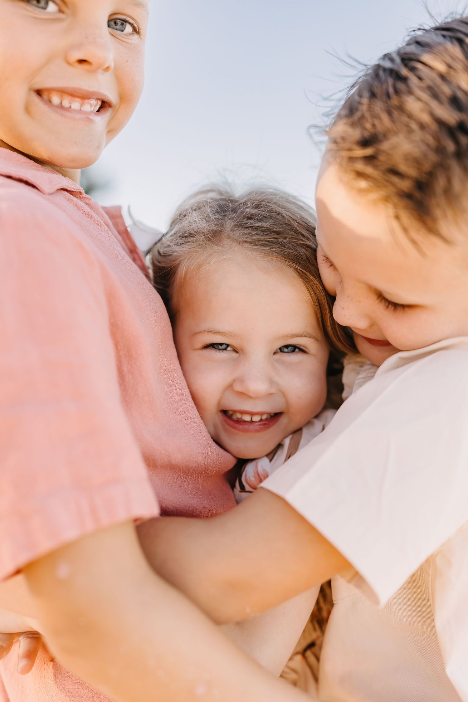 Three people share a joyful close embrace while smiling and laughing together in warm sunlight.