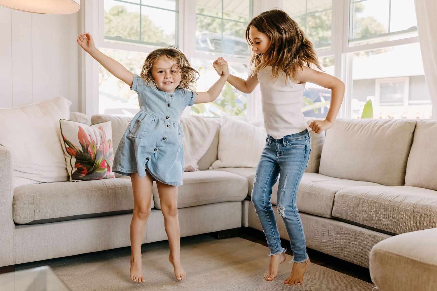 Two people dancing and jumping together in a bright living room with a beige sectional sofa.