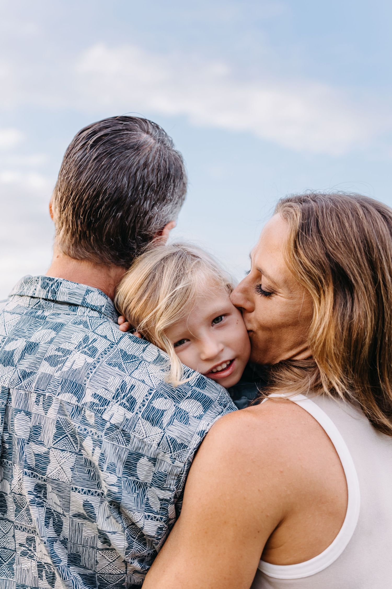 A close family moment captured outdoors against a blue sky with white clouds.
