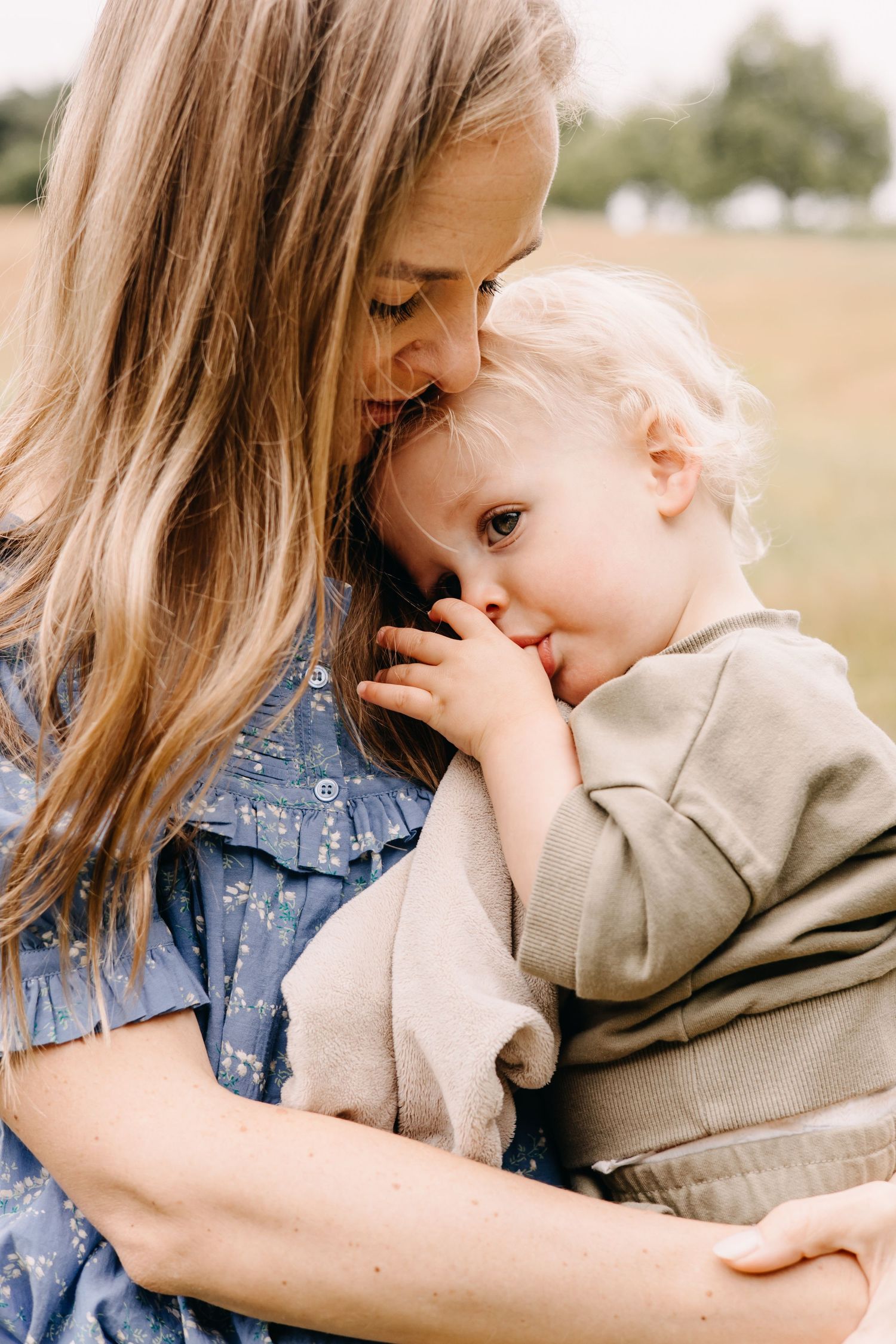 A tender moment of closeness as a baby in beige sweater cuddles against their caregiver in a blue dress outdoors.