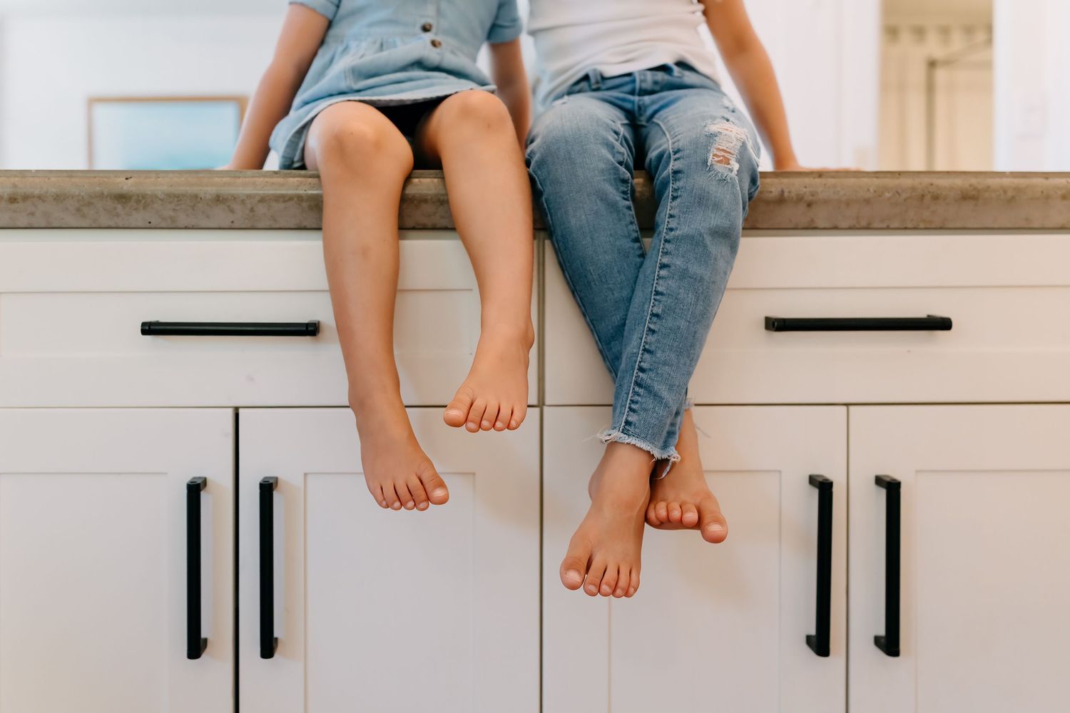 Two people sitting casually on a white kitchen counter wearing shorts and jeans with bare feet dangling down.