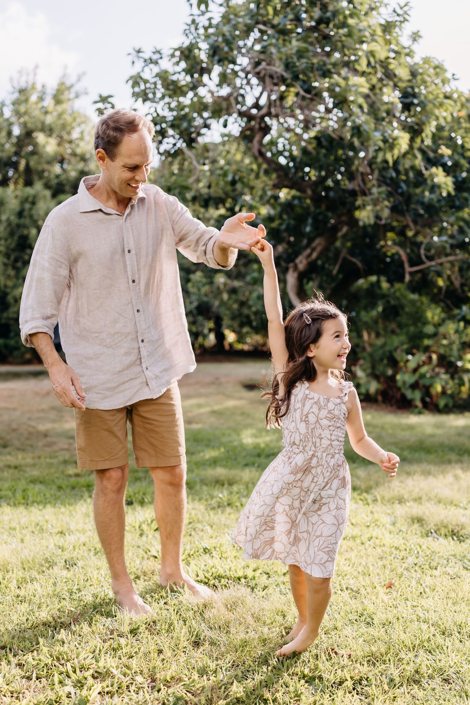 Joyful parent and young child dancing and playing together on a sunny lawn with green trees in the background.
