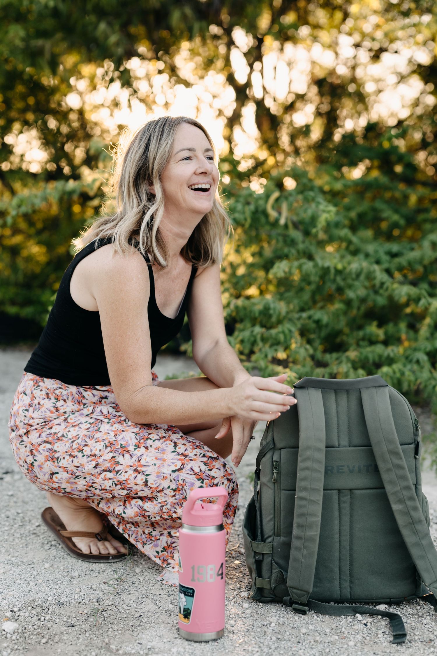 Outdoor portrait showing joyful laughter while crouched near a backpack and pink water bottle at golden hour.