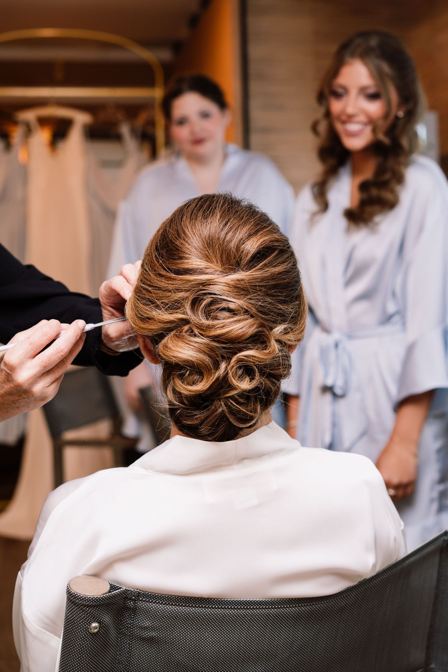 Elegant updo hairstyle being styled for wedding preparation while others in white robes look on in background.