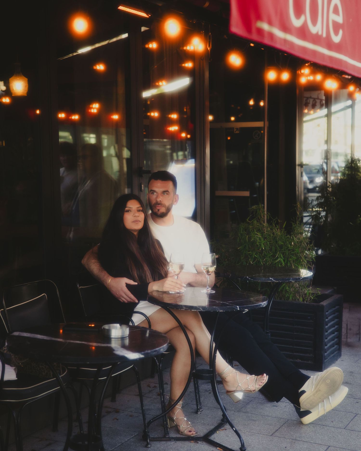 A couple enjoys drinks at an outdoor cafe patio at night with warm lighting and planters creating an intimate atmosphere.