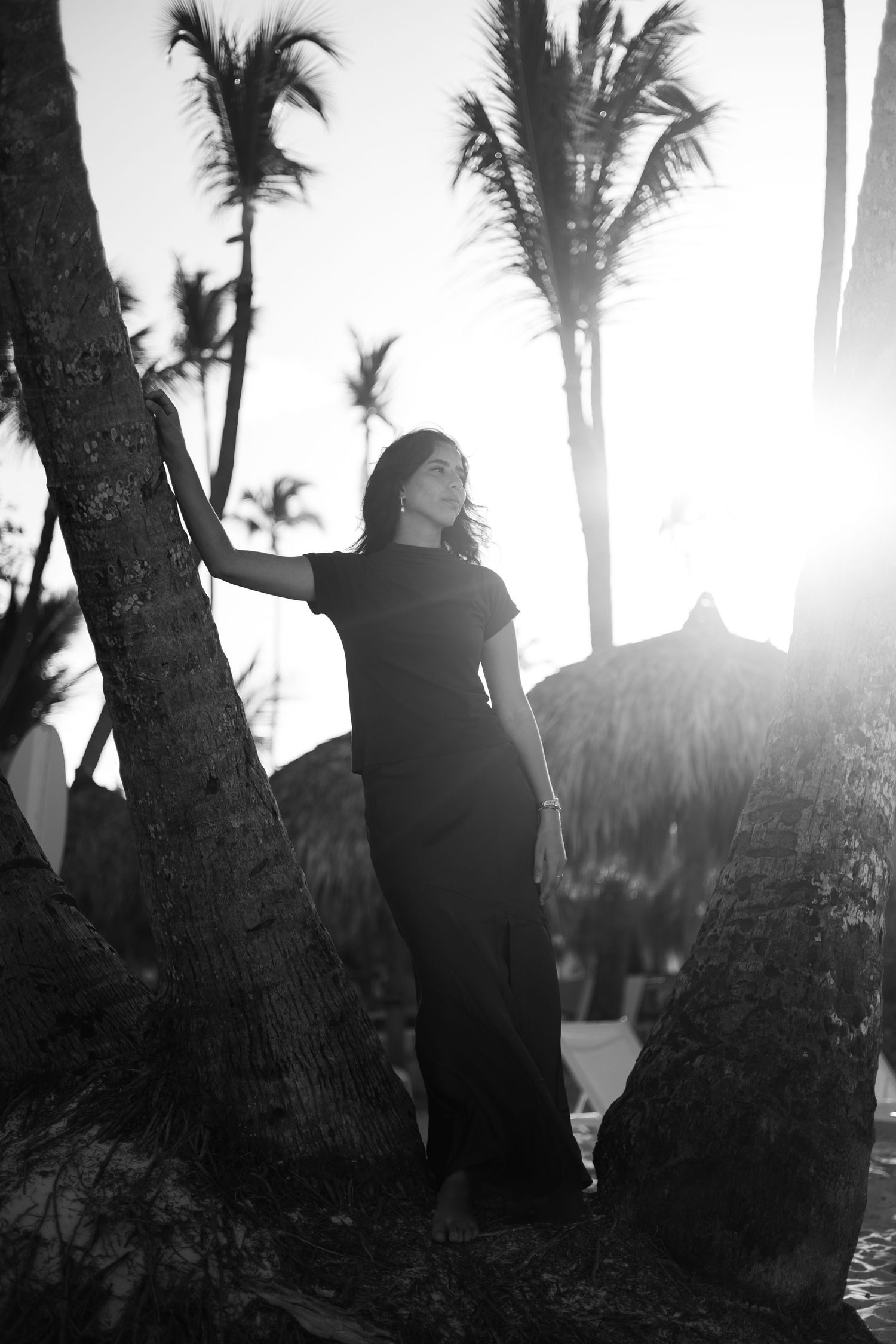 Silhouette of woman in long dress posing between palm trees at tropical sunset.