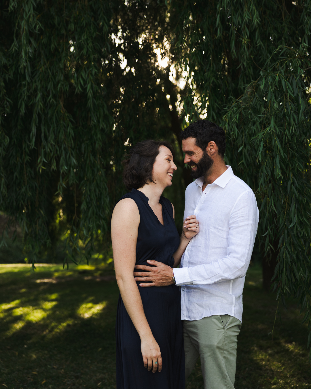 A romantic couple shares an intimate moment beneath drooping willow branches in a sunlit garden setting.