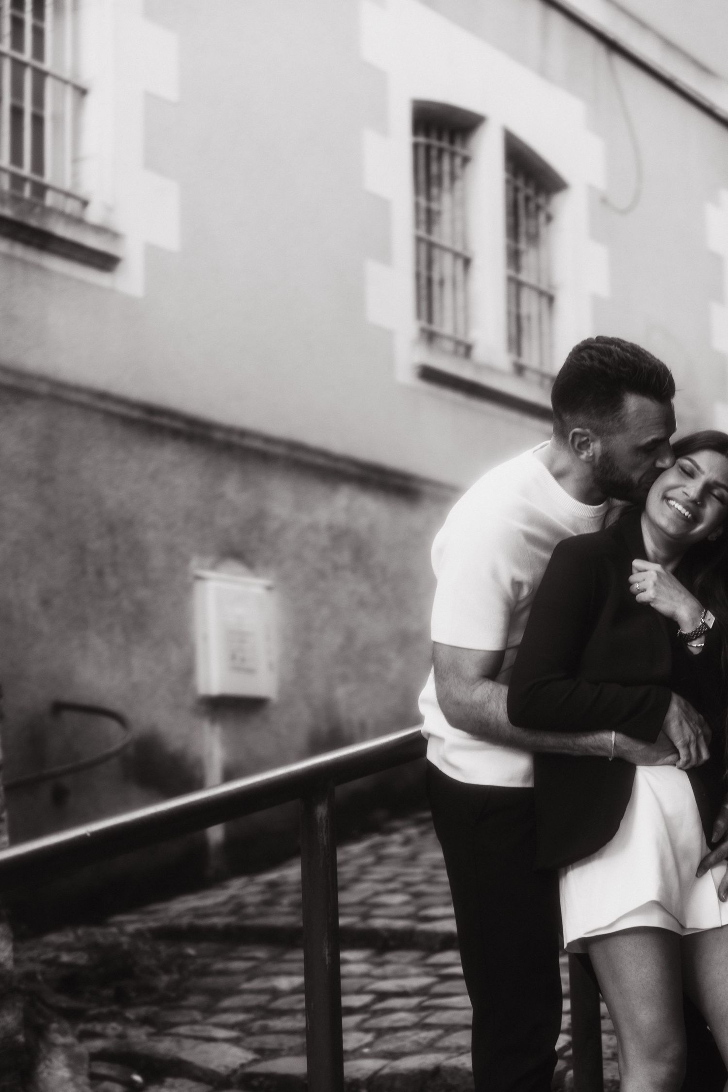 A romantic couple embraces and laughs together on cobblestone steps in black and white photography.