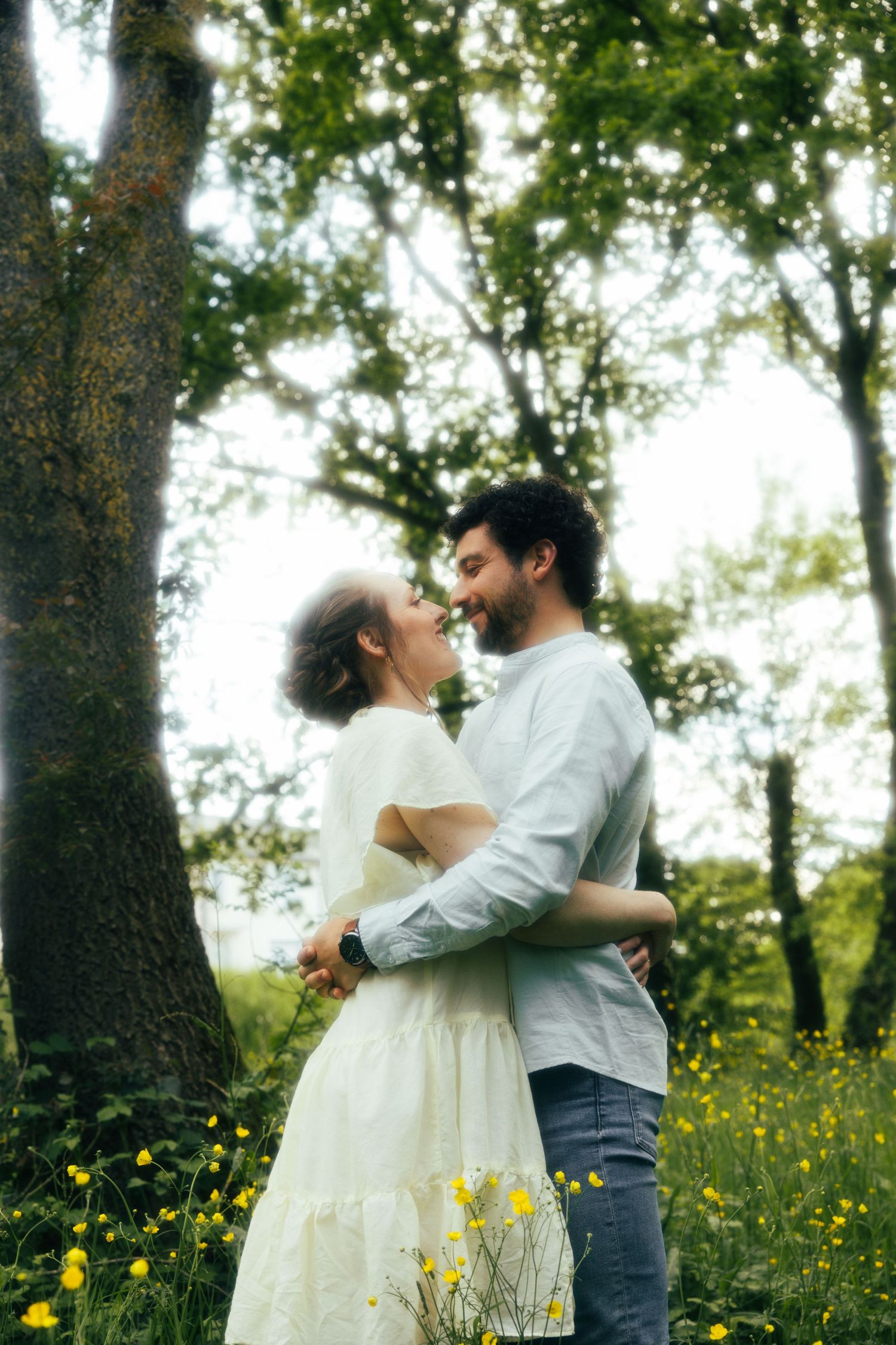 A romantic couple embraces in a spring meadow with yellow wildflowers and trees in soft dreamy lighting.