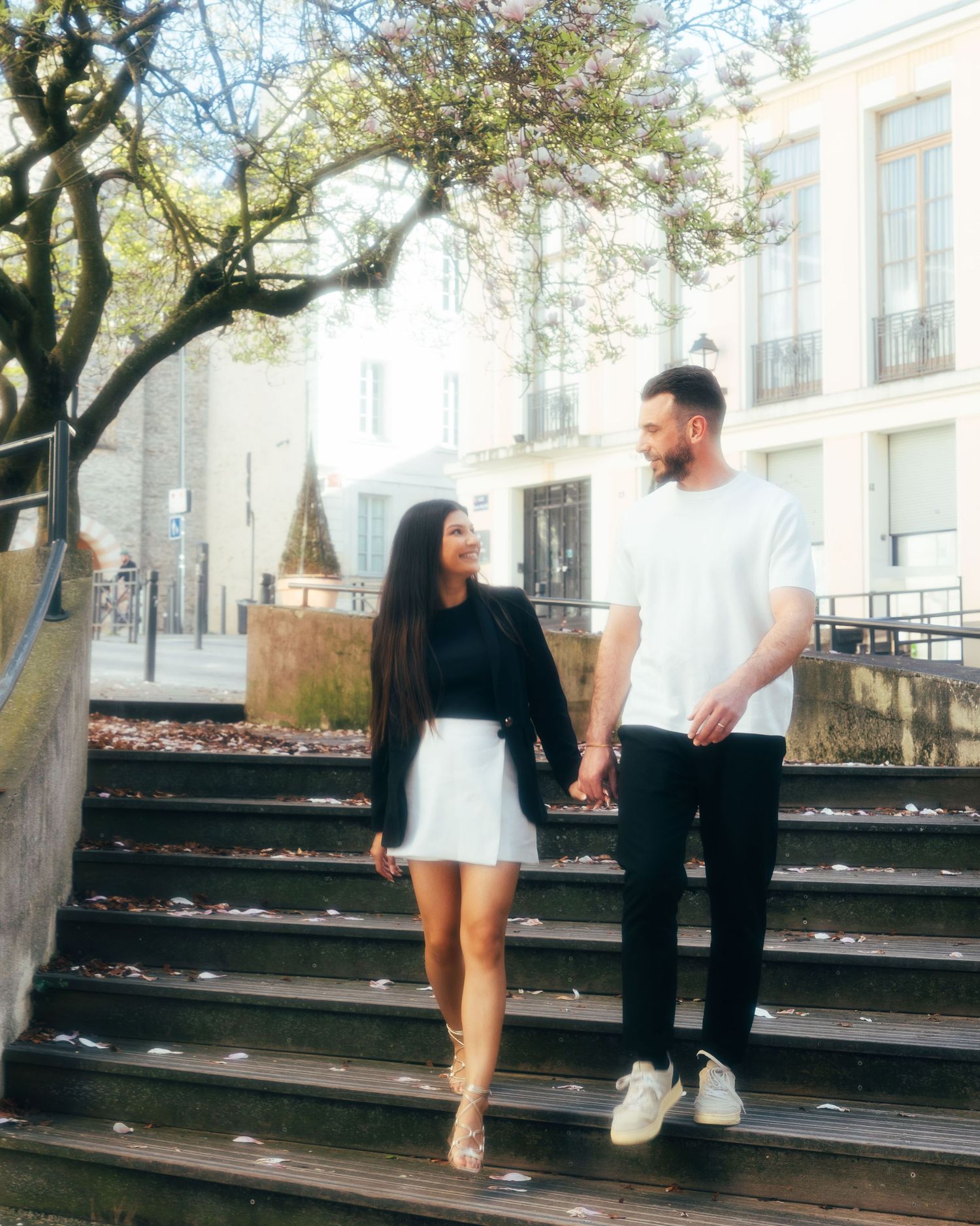 A couple standing on outdoor stone steps, holding hands against an urban backdrop on a sunny day.