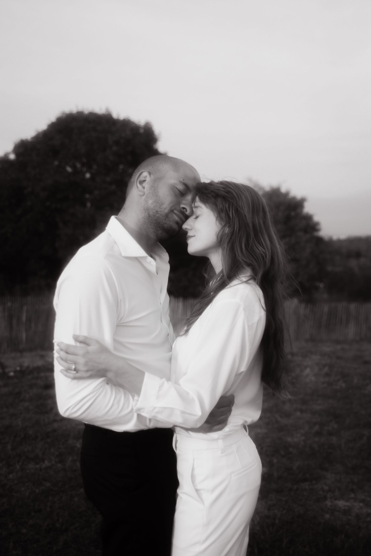 An intimate black and white portrait captures a romantic couple in white clothing embracing outdoors.