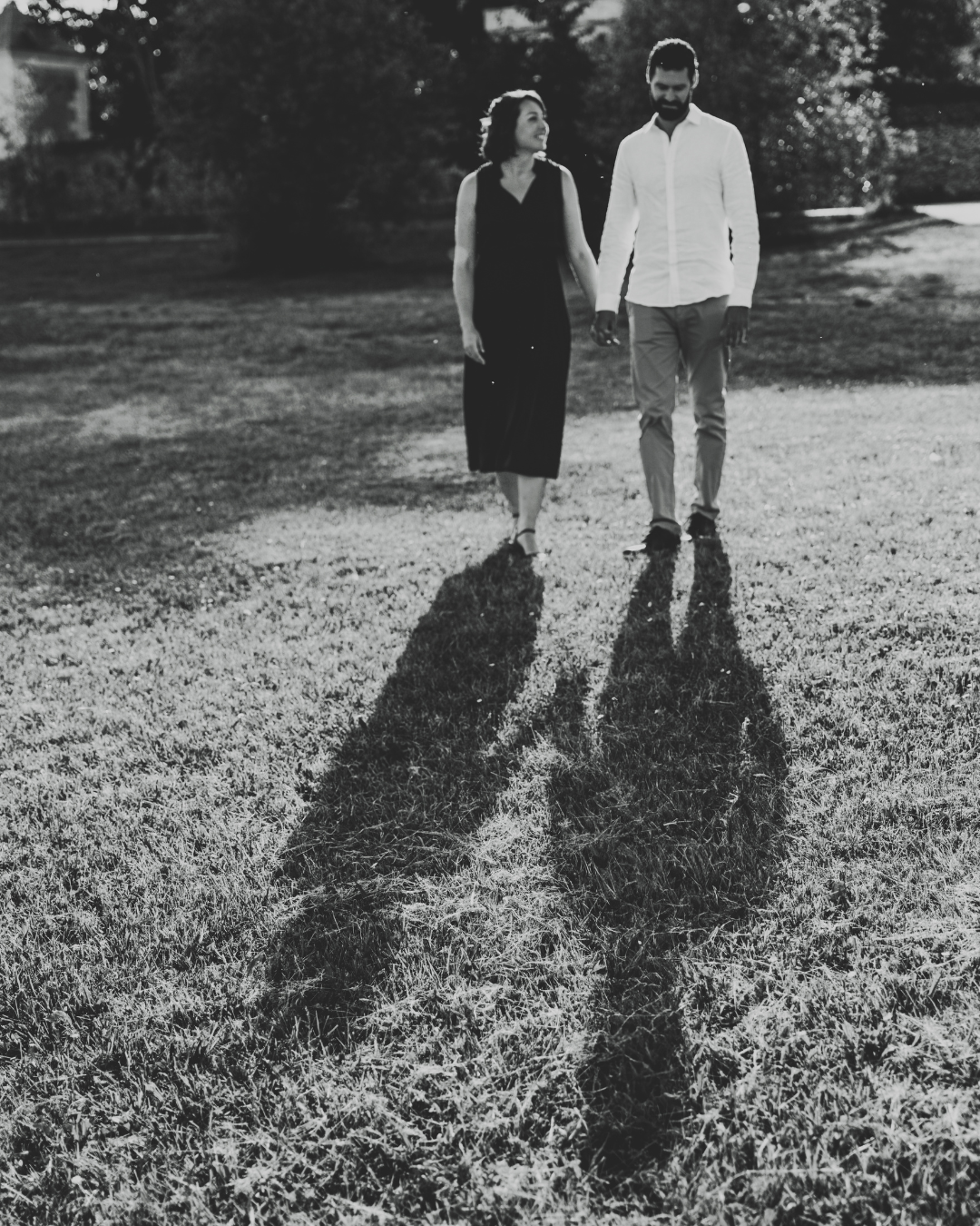 A couple stands together on a grassy lawn with their dramatic long shadows cast behind them in black and white.