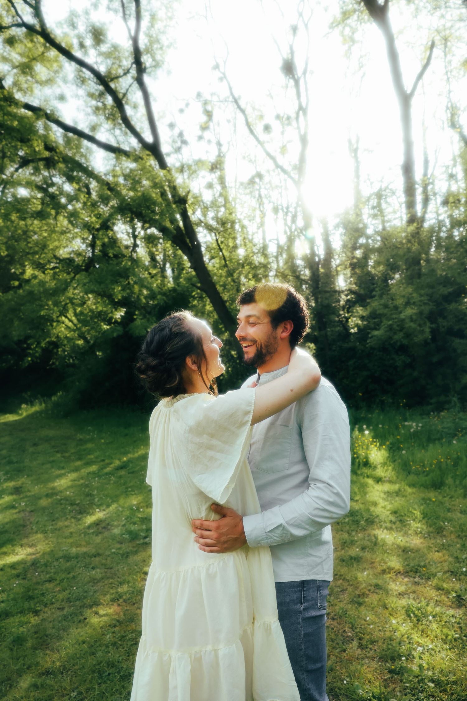 A couple embraces in a romantic moment outdoors in a sunlit garden surrounded by lush green trees.