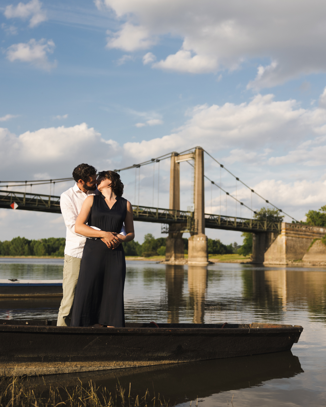 A couple shares an intimate moment on a dock by a suspension bridge over a river during golden hour.
