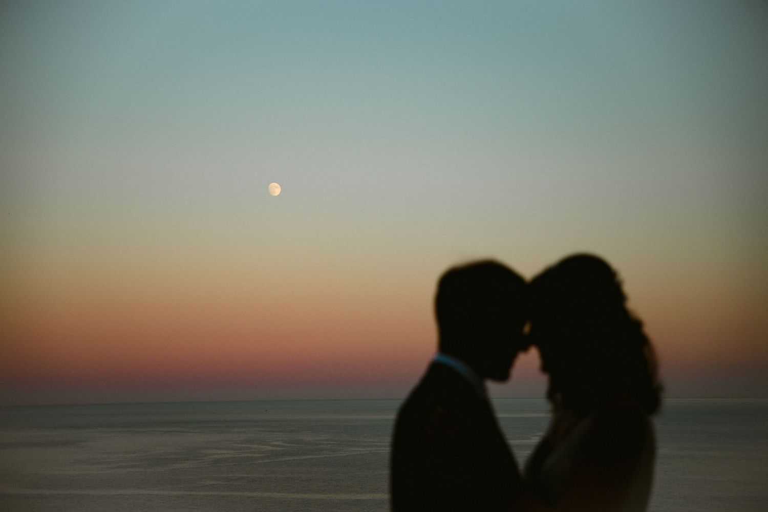 Silhouetted romantic couple embracing at sunset against an ocean backdrop with a glowing moon in the sky.