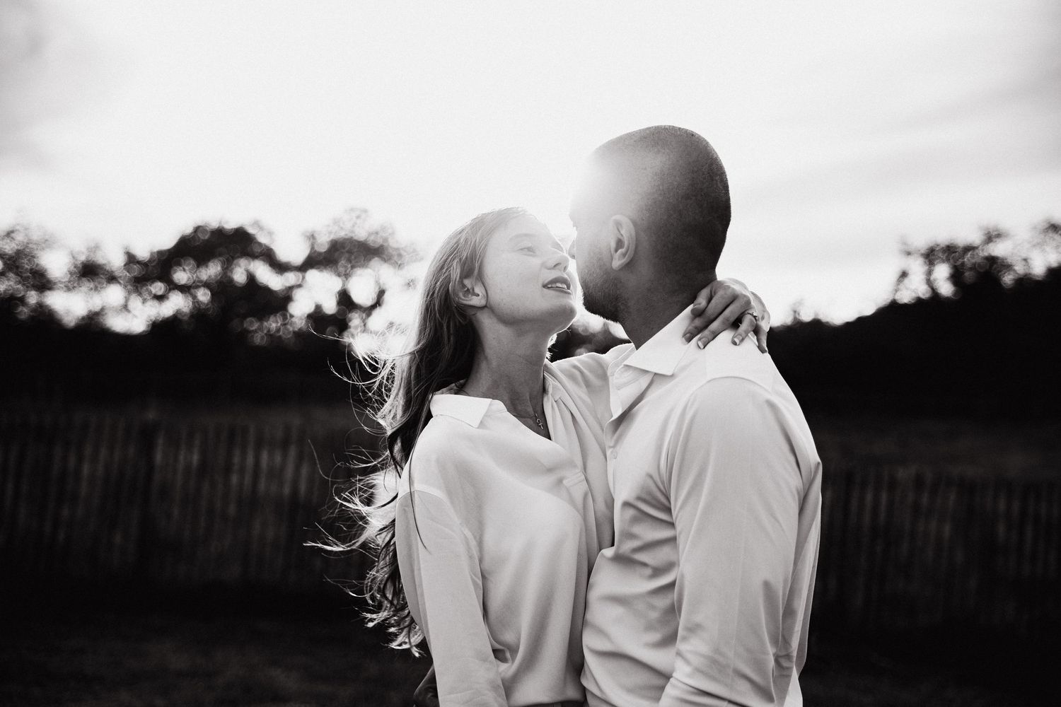 Two people share an intimate romantic moment at sunset with a fence in the background.