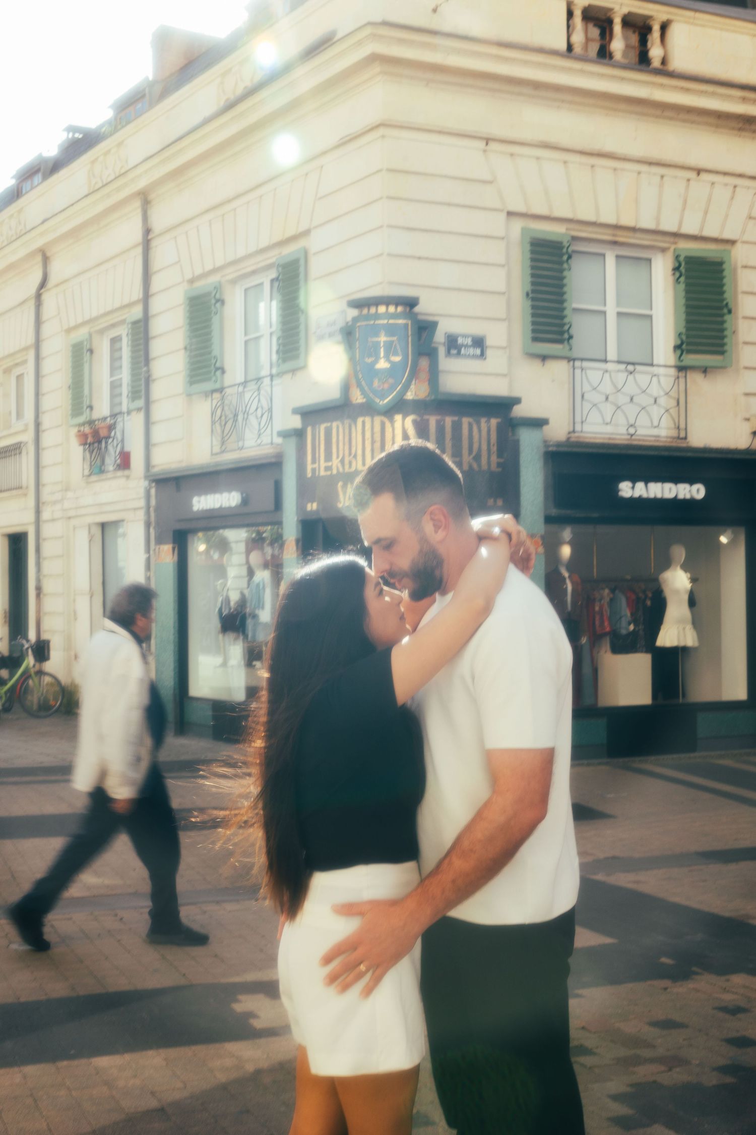 A couple shares an intimate moment on a European street corner with vintage storefronts and green shuttered windows.