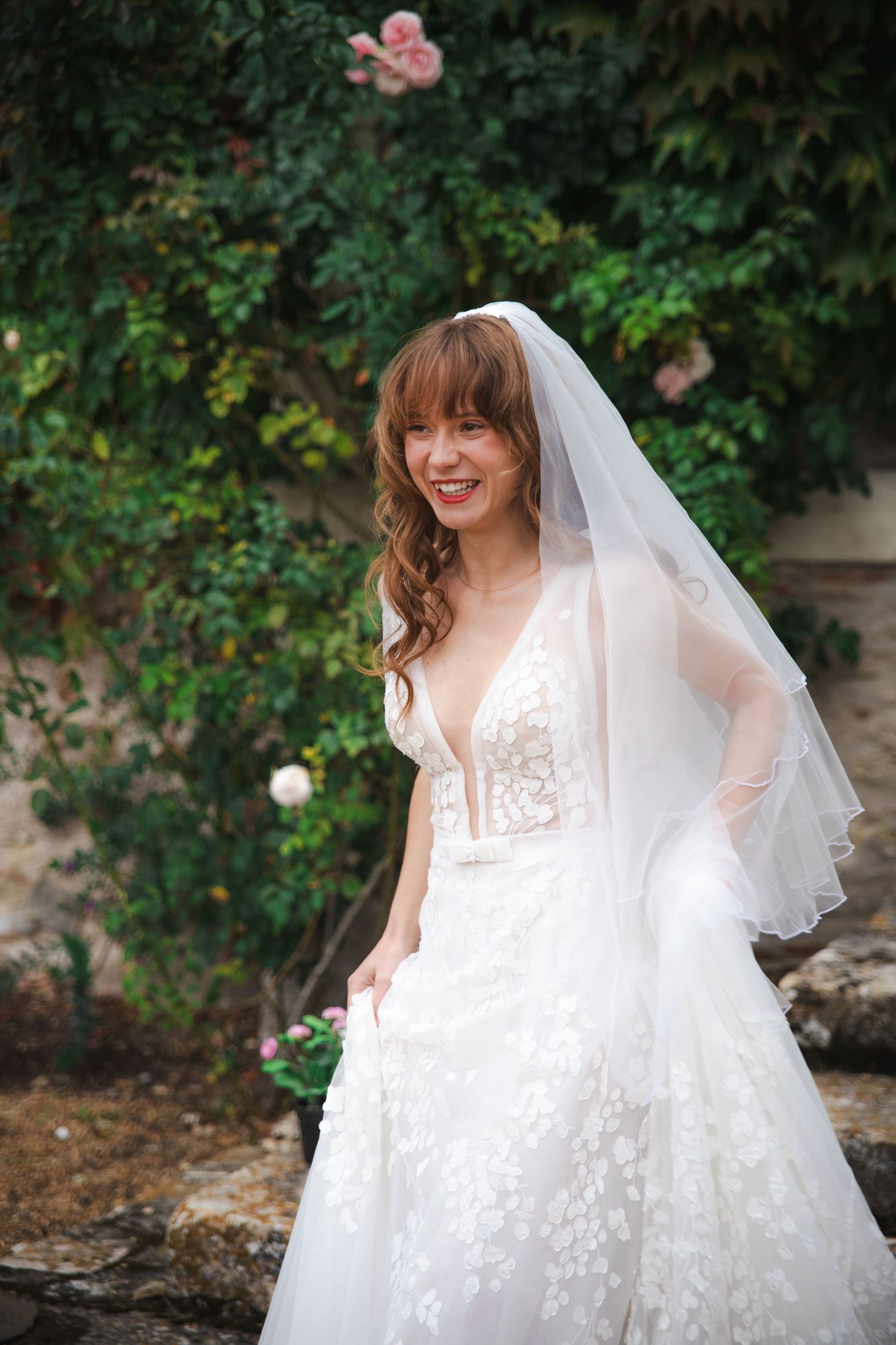 A bride wearing a beautiful white lace wedding gown with a flowy veil poses in front of a garden with roses.
