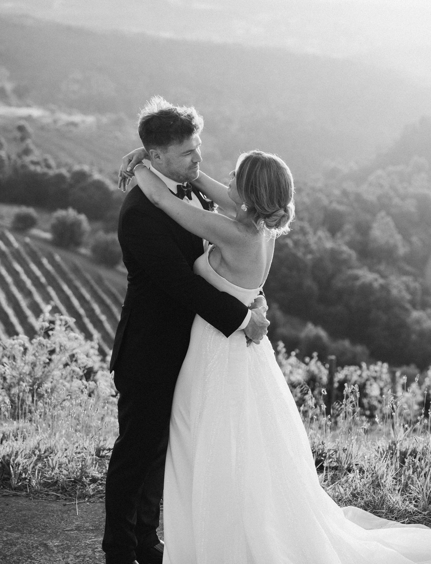 Black and white photo of newlyweds embracing on a hillside overlooking vineyards in Stellenbosch, South Africa.