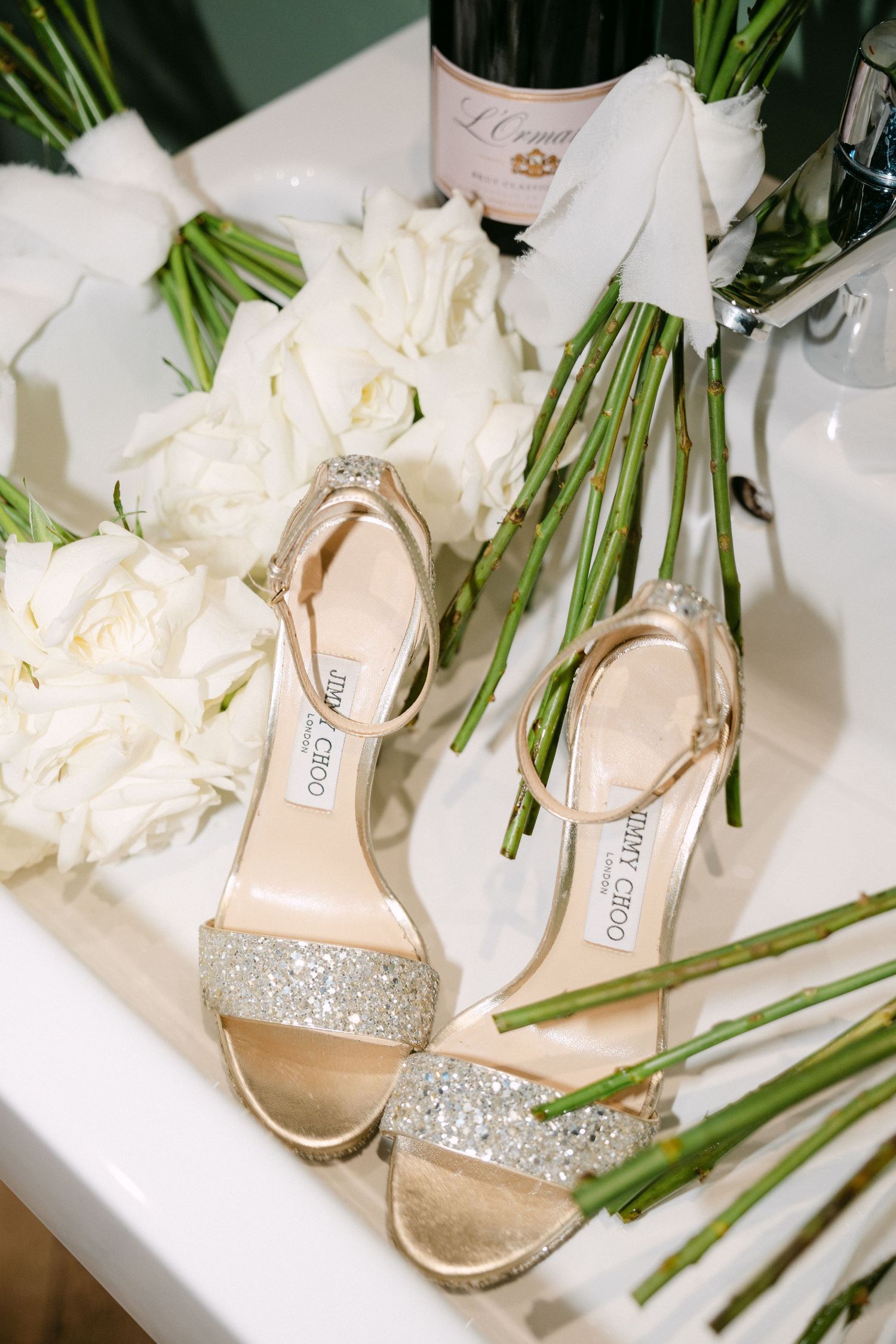 Glittery Jimmy Choo bridal sandals displayed with white roses and green stems.