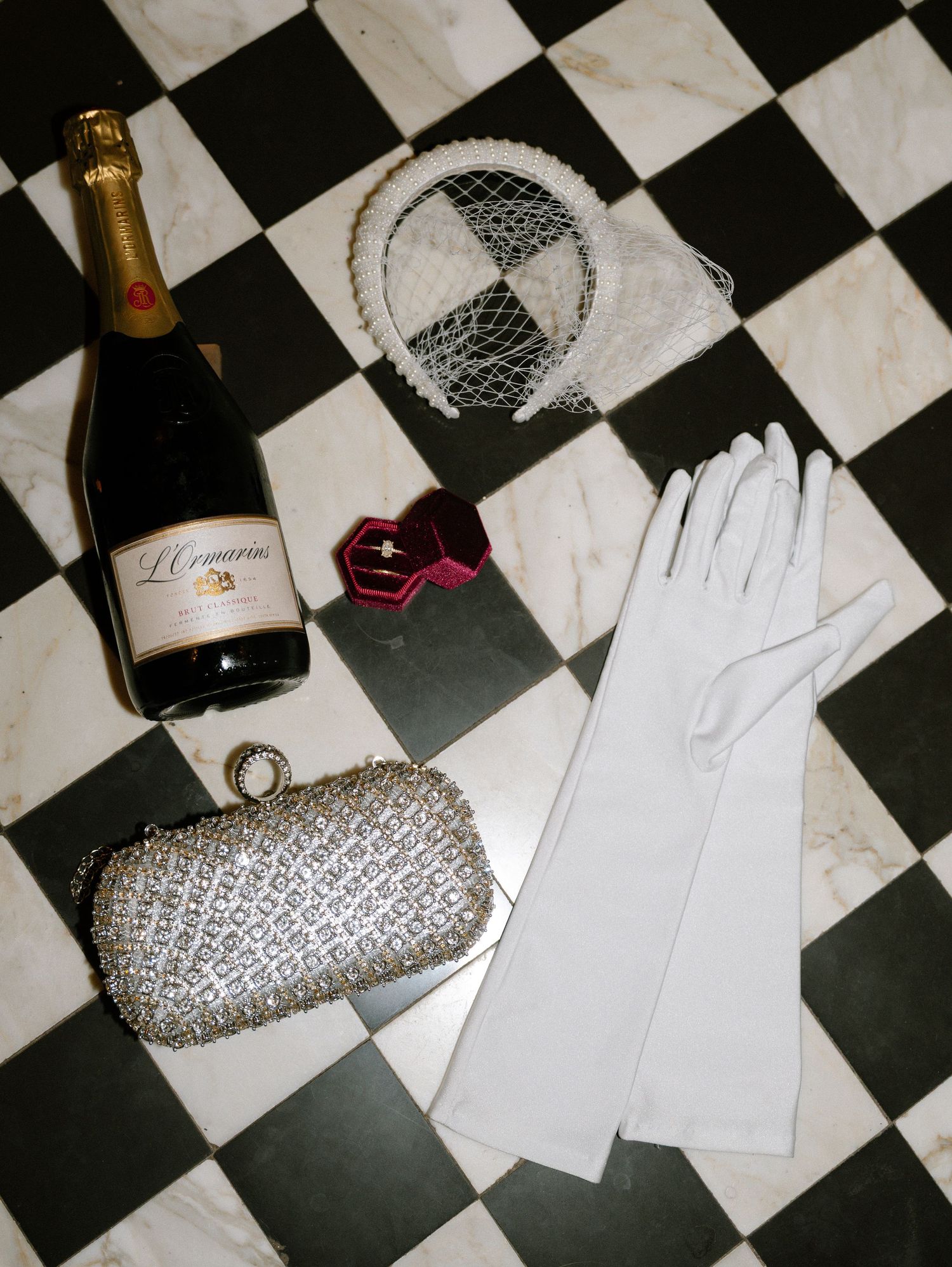 Elegant wedding accessories including champagne, white gloves, and a silver clutch on a checkered floor.