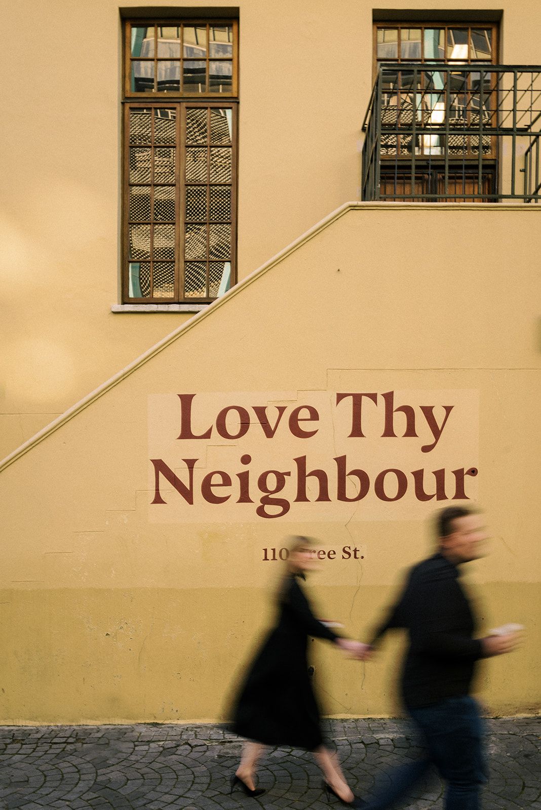 Wall with 'Love Thy Neighbour' text and blurred figures walking past.