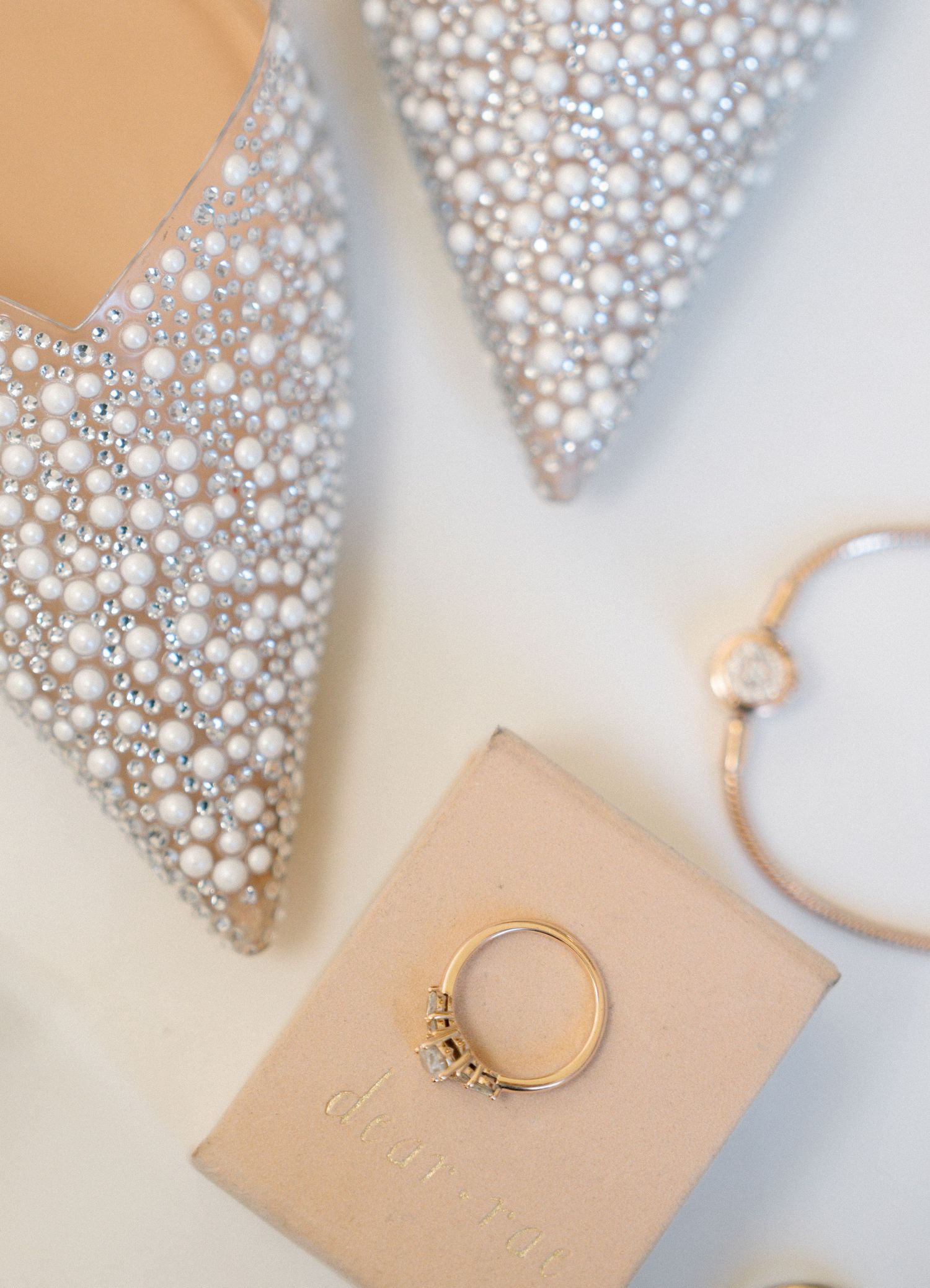 Pearl-encrusted wedding shoes and engagement ring displayed on blush colored surface.