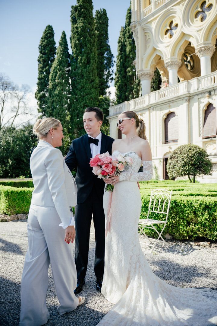 A wedding ceremony takes place on a sunny day in front of an ornate building with cypress trees and manicured gardens.