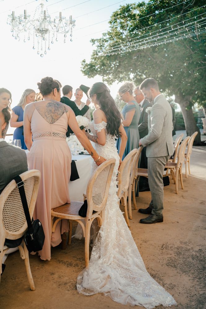 Wedding guests gather around a long dining table under string lights and chandeliers.