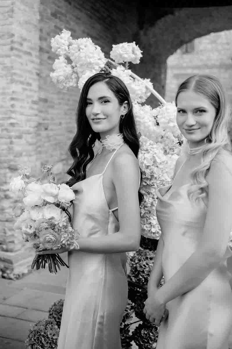 Black and white portrait of attendants in elegant dresses holding bouquets against stone backdrop.