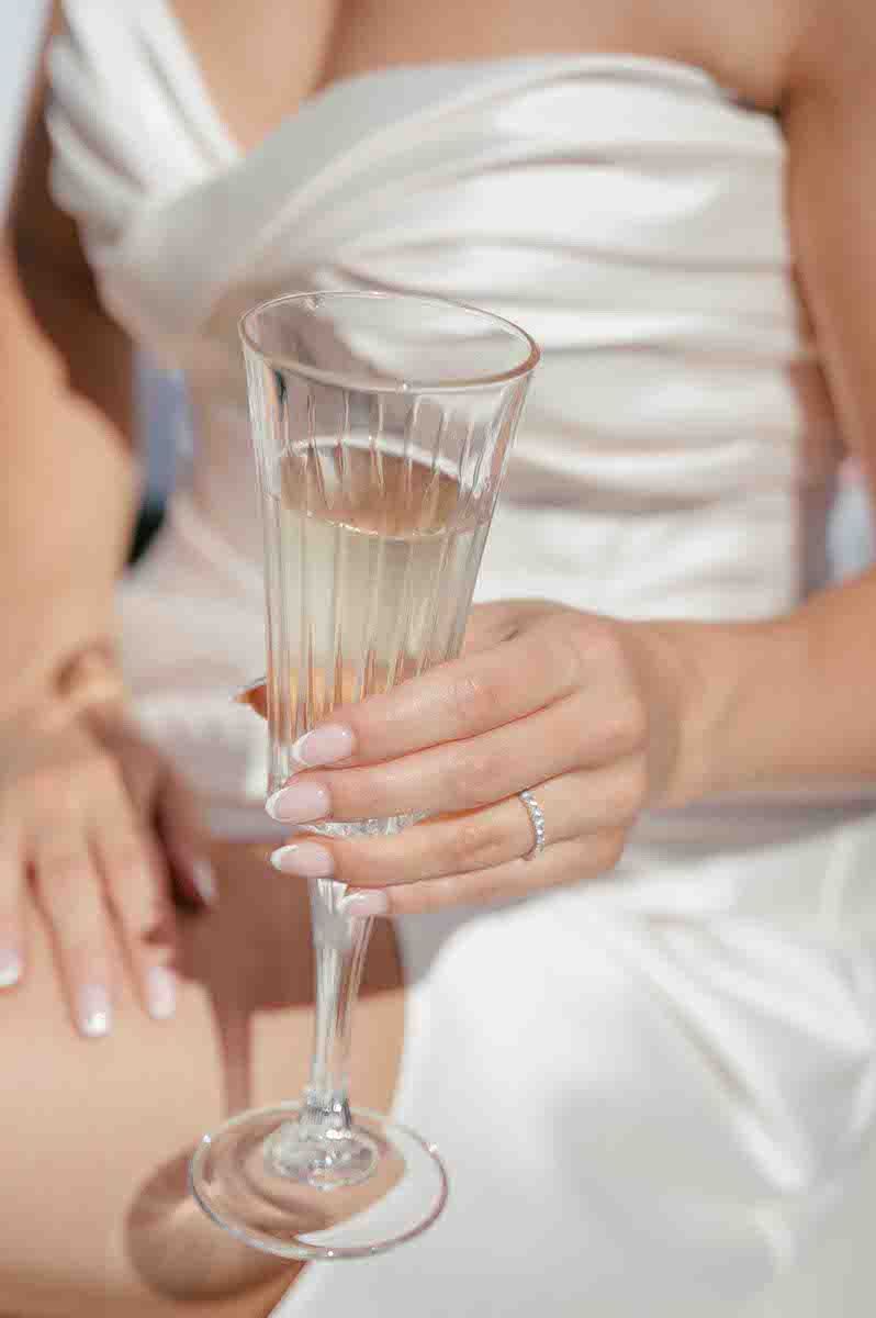 Close-up of champagne glass held by manicured hand with wedding ring and white dress.