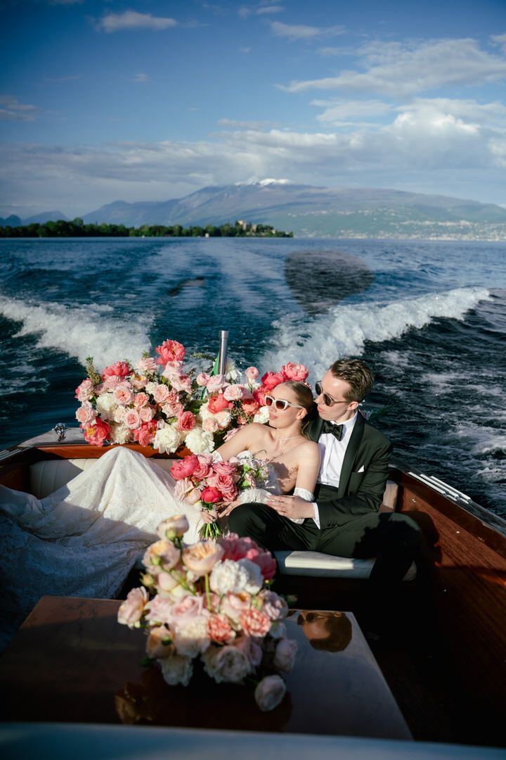 A couple on a boat with pink and white floral arrangements cruising across stunning blue waters near a mountainous coastline.