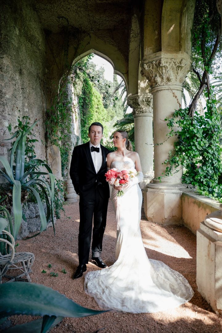 A couple poses in formal wedding attire under an elegant stone archway with climbing vines and natural greenery.