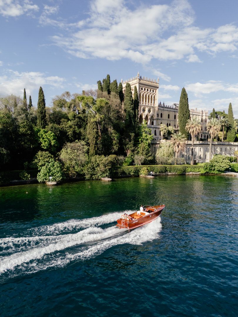 A wooden motorboat cruises across blue waters near a historic villa with cypress trees on Lake Garda in Italy.