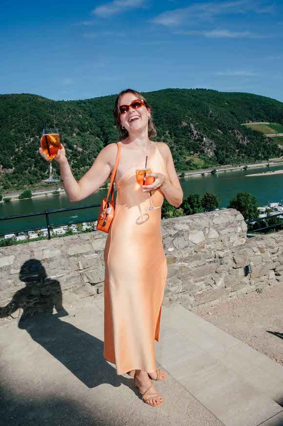A person in a peach colored dress holds drinks while standing on a stone wall overlooking the Rhine River valley.