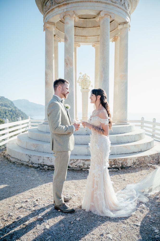 Couple shares intimate moment under classical stone gazebo with mountain backdrop.