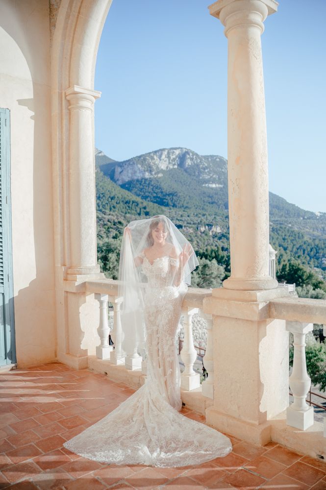 A bride in a lace wedding dress poses on a Mediterranean terrace with mountain views.