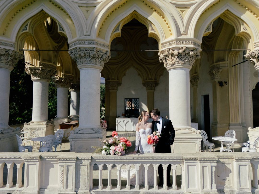 A couple stands on an elegant stone balcony with ornate columns and Gothic arches on a sunny day.