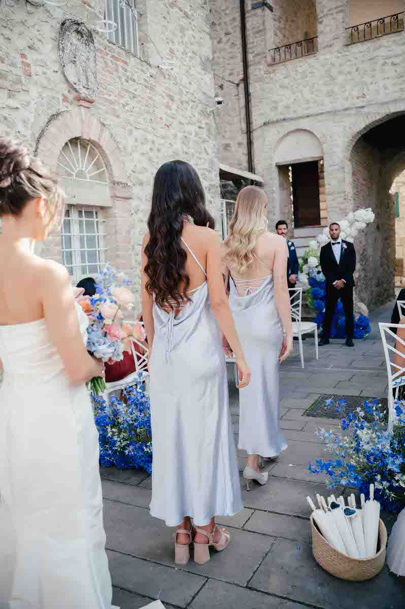 Wedding party stands in front of a stone castle wall with blue floral decorations and elegant white chairs.