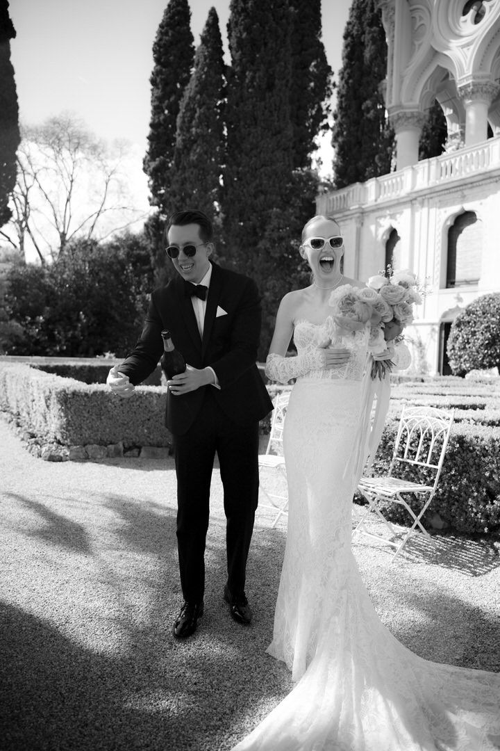 A bride in a fitted white gown and groom in classic tuxedo pose together on a garden terrace with cypress trees.