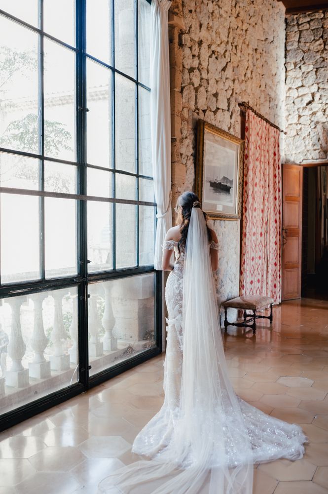 Bride in flowing white lace gown with long train stands elegantly by large window in rustic stone interior.