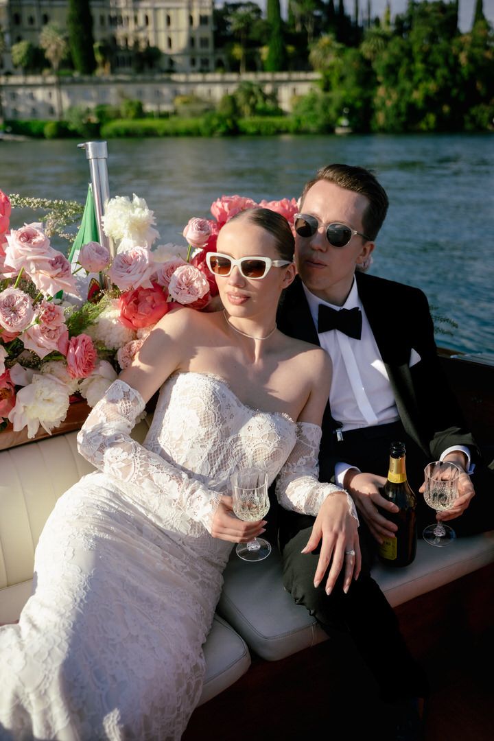 A stylish couple in sunglasses relaxes on a boat with champagne and pink flowers beside a waterfront estate.