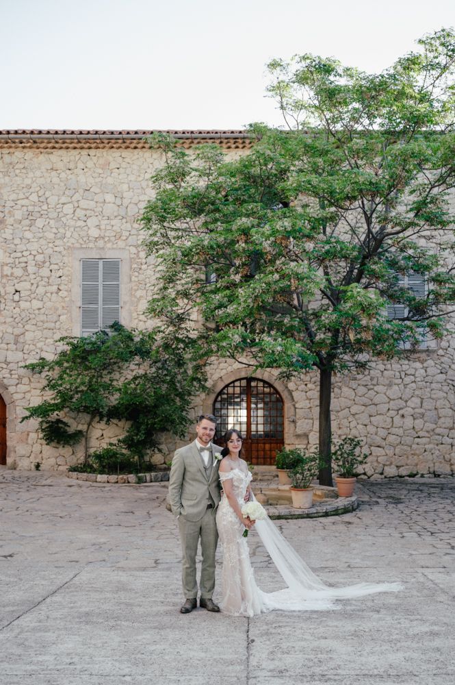 A wedding couple stands in front of a rustic stone building with an elegant arched doorway and green tree.