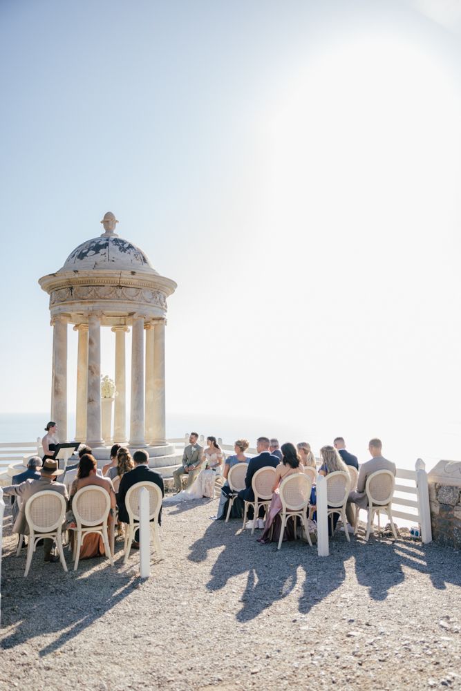 Wedding guests seated at elegant ceremony under Mediterranean-style gazebo overlooking ocean vista.
