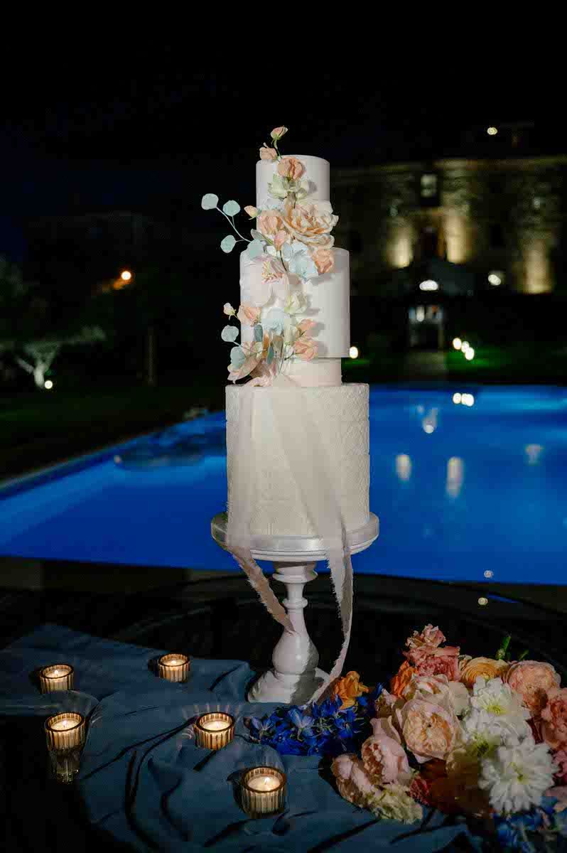 Elegant white wedding cake with peach flowers displayed by illuminated pool at night.