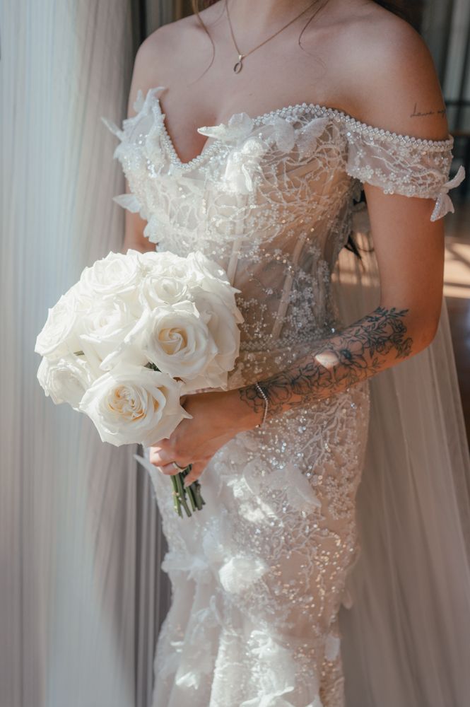 Close-up detail of beaded off-shoulder wedding dress with white rose bouquet.