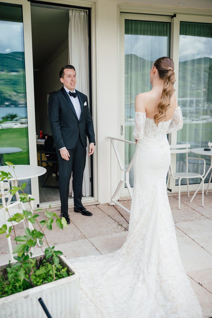 First look wedding moment as groom in black tuxedo sees bride in lace mermaid dress on scenic mountain balcony.